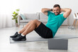 © Prostock-studio - Cheerful young muscular caucasian guy doing abs exercises and looking at computer in living room interior with bottle of water. Sports training, coach remotely, new normal during covid-19 pandemic