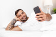 © Prostock-studio - Smiling millennial attractive guy with beard lies on white bed, chatting and reads news on smartphone in bedroom interior. Rest and relax at home, new application, great offer, ad and social networks