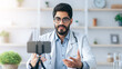 © Prostock-studio - Medical blog, webinar and online consultation during covid-19 social distancing. Friendly 30s arabian doctor in coat and glasses, gesturing and looking at phone webcam at table in office interior