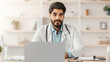© Prostock-studio - Visit to doctor remotely, online consultation, telemedicine during COVID-19 pandemic. Young arabian bearded guy in white coat with stethoscope sits at table in office interior in clinic, copy space