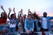© pressmaster - Group of multiethnic young adults dancing and enjoying rooftop party while Caucasian man DJ operating mixing console, people raising hands and smiling, cityscape visible in background