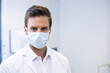 © wavebreak3 - White male healthcare worker standing in clinic wearing lab coat and mask with dispenser, monitor