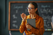 © Seventyfour - Caucasian young adult woman standing in classroom holding digital tablet, gesturing while explaining lesson content, blackboard with handwritten notes visible in background, language school setting