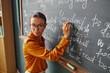 © Seventyfour - Caucasian young adult woman writing English phrases on chalkboard, wearing glasses, standing in classroom, teaching language lesson, looking over shoulder toward students
