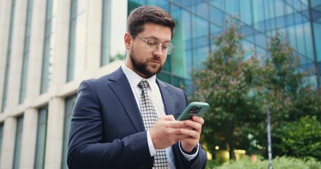 Confident Caucasian man in jacket and tie walking outdoors on a sunny summer day, using a smartphone and typing a message with a modern glass building behind him