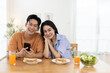 © eakgrungenerd - Smiling Asian couple enjoying morning together at breakfast table, holding smartphone. Concept of modern relationship, digital lifestyle, love and comfort in cozy home atmosphere. looking at camera.