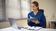 © matinee - Smiling female healthcare worker in blue scrubs uses a digital tablet and laptop during an online medical session.