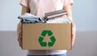 © otra - A woman holds a cardboard box filled with used clothes and books for donation, featuring a green recycling symbol for sustainable reuse