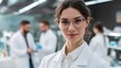 © Harry - Confident female scientist with glasses smiles warmly in a modern laboratory setting, colleagues blurred in background working diligently