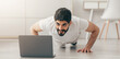 © Prostock-studio - Training with coach remotely, modern technology for social distancing and sports at home. Happy arabian bearded millennial male push-ups, looks at laptop with dumbbells, bottle of water in living room