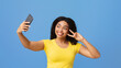 © Prostock-studio - Excited african american lady taking selfie with smartphone and showing victory gesture on blue studio background. Happy black woman making photo of herself on mobile phone, having fun and fooling