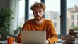 © miss irine - Young man with curly red hair, beard works intently on laptop at home office. Sits at desk wearing orange sweater, focused expression, calm demeanor. Window view provides city backdrop, technology,