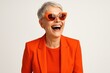 © Jelena - Stylish senior woman in vibrant orange monochrome outfit and cool sunglasses laughing confidently modern age positive aging fashion studio portrait on solid backdrop