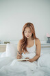 © WMSTUDIO - Young woman with long hair sitting on bed in white comfortable bedding, writing in notebook for personal reflection or journaling in peaceful morning light