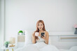© WMSTUDIO - Young woman relaxing in bed with a smartphone, holding a cup of coffee, enjoying her morning routine in a bright and cozy bedroom setting