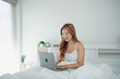 © WMSTUDIO - Young woman working on laptop in cozy bedroom, smiling while sitting on bed with white sheets, relaxed atmosphere, natural light from window