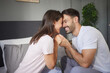 © Stockphotodirectors - A couple is playfully holding hands and smiling at each other while sitting on their bed in a comfortable room, radiating happiness and affection on a sunny morning.