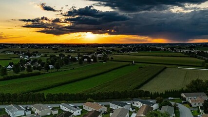 Wall Mural - A hyper lapse of a golden sunlight fades as the sun sets behind rolling hills and vibrant green fields. The peaceful rural landscape features farmland, trees, and distant homes under a colorful sky.