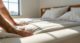 A man's hands neatly making a bed and smoothing a white duvet in a bright, sun-drenched bedroom