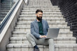 Portrait of cheerful businessman sitting on staircase at financial district with laptop and typing on it.