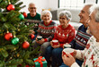 © Patrick - Senior friends in festive sweaters enjoying hot drinks and conversation by Christmas tree with decorations and presents in cozy living room during holiday season