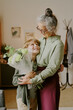 © AnnaStills - Caucasian senior woman embracing Caucasian boy while holding bouquet of flowers, both smiling and making eye contact, standing together in home setting, showing affectionate moment