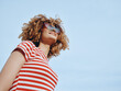 © SHOTPRIME STUDIO - Portrait of a joyful young person with curly hair, wearing a red and white striped shirt and sunglasses, standing outdoors against a clear blue sky.