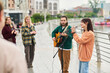 © pressmaster - Young Caucasian man playing acoustic guitar and young Caucasian woman singing into microphone performing outdoors on riverside walkway