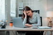 © Crystal - Asian office worker sitting at her desk, struggling with a severe headache while using her laptop, feeling overwhelmed and stressed