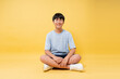 © Q88 - Smiling teen sits cross-legged on a bright yellow background wearing casual clothing and sneakers