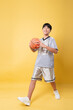 © Q88 - Teen basketball player in uniform smiles holding ball against a plain yellow background Full length