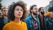 © serdon - Young woman with curly hair and glasses looking up with hope while standing in a diverse crowd at an outdoor event