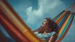 © serdon - Young woman with curly hair relaxing with eyes closed in a colorful striped hammock on a sunny day under a clear blue sky