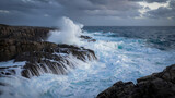 Powerful ocean waves crash against rugged, dark rocks under a dramatic, cloudy sky, creating a dynamic display of natures force