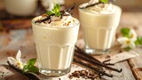 Two glasses of creamy vanilla milkshake, topped with whipped cream, cocoa powder, and vanilla beans, sit on a rustic wooden surface, creating a delicious and inviting image.