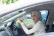 © eakgrungenerd - Confident Muslim woman wearing hijab sitting in car driver's seat, holding steering wheel and smiling, symbolizing independence, driving skills and modern lifestyle.