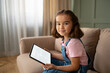 © MSTHUMAYRAAKTER - Smiling girl reading e-book on tablet, sitting on sofa, casual home setting, happy child, digital learning, education concept.