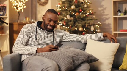 Young handsome African man sitting on sofa with different pillows near beautiful New Year s tree. Guy chatting, using smartphone. Evening. Indoor.