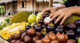 Hands selecting a fresh mangosteen from a vibrant display of tropical fruits like salak and jackfruit