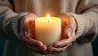 © momostudio - A lit candle held gently in cupped hands against a neutral background.