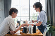© whyframeshot - Asian mature man enjoys morning coffee at cafe using drip coffee bag in beige cup on white wood table, while young adult friends relax making specialty coffee as hobby activity at home