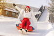 © Vladimir Razgulyaev - A young woman with a bright red beanie and matching leggings is joyfully tubing down a snowy slope. Dressed in a cozy white coat, she throws her arms wide open, embracing the thrill of winter sports.