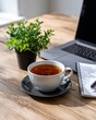© Николай Срибяник - Cozy home office workspace featuring hot tea in white cup, laptop computer, green plant, and spiral notebook on natural wood desk surface