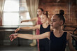 © DragonImages - Caucasian teenage girls practicing ballet positions at barre in dance studio with female instructor guiding in background, focused expressions, hair in buns
