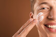 © New Africa - Handsome young man applying cream onto his face on brown background, closeup. Space for text