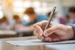 © Wa Bale - Close-up of a hand holding a pen, writing on paper in a blurred classroom setting.  The image conveys focus, concentration, and the act of learning or testing