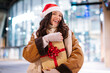 © Home-stock - Happy woman in Santa hat embracing gift box, buying Christmas present, preparing for winter holidays, standing in front of shopping mall in the evening
