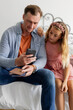 © wavebreak3 - Father and daughter sitting on bed, looking at smartphone together, smiling