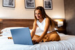 © Zoran Zeremski - Female entrepreneur working from hotel room sitting on bed and using laptop.