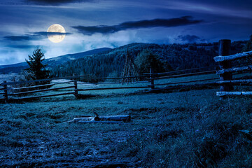 Wall Mural - mountainous rural landscape in autumn at night. wooden fence near the path through grassy meadow on the hillside. scenery with fir forest on the mountains in full moon light under sky with clouds